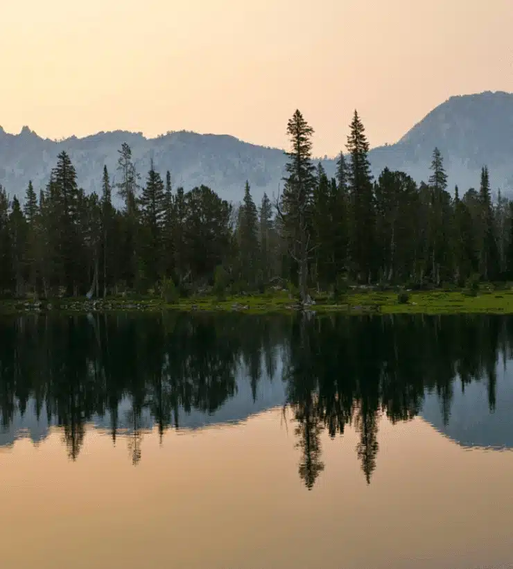 A calm lake reflecting pine trees and mountain silhouettes during twilight at Sawtooth National Recreation Area, Idaho.